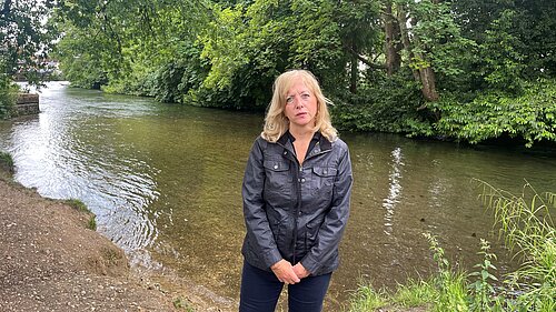 Liz Jarvis MP standing beside a river, wearing a black jacket and wellington boots. She is surrounded by trees and greenery, with a serious expression, highlighting concern for the local environment.